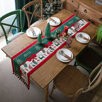La table est joliment décorée pour les fêtes, avec une nappe festive, de petits arbres verts et des décorations rouges, le tout placé sur une surface en bois clair.