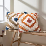 Coussin décoratif avec motif géométrique coloré et franges, placé sur un banc en bois dans un intérieur lumineux.