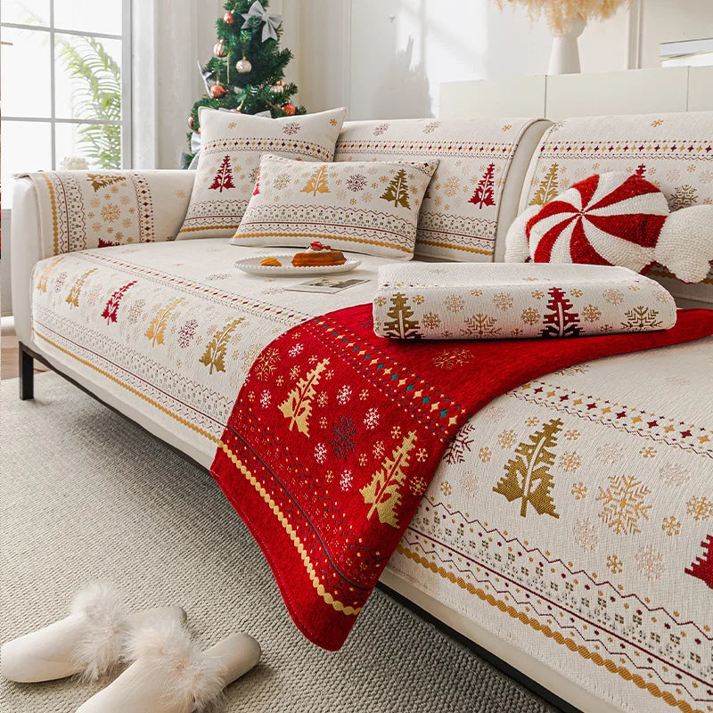 L'image montre un ensemble de coussins, de couvertures et de décorations avec des motifs de flocons de neige et d'arbres de Noël dans des tons de rouge, d'or et de beige, créant une ambiance chaleureuse et festive dans une pièce illuminée par une fenêtre.