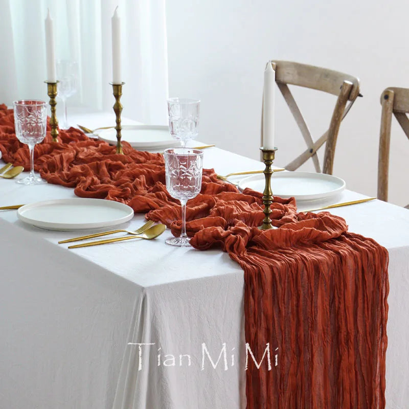 Une table élégante avec une nappe blanche, des bougies, des verres de cristal et une décoration de tissu froissé en couleur rouille, créant une atmosphère chaleureuse et raffinée.