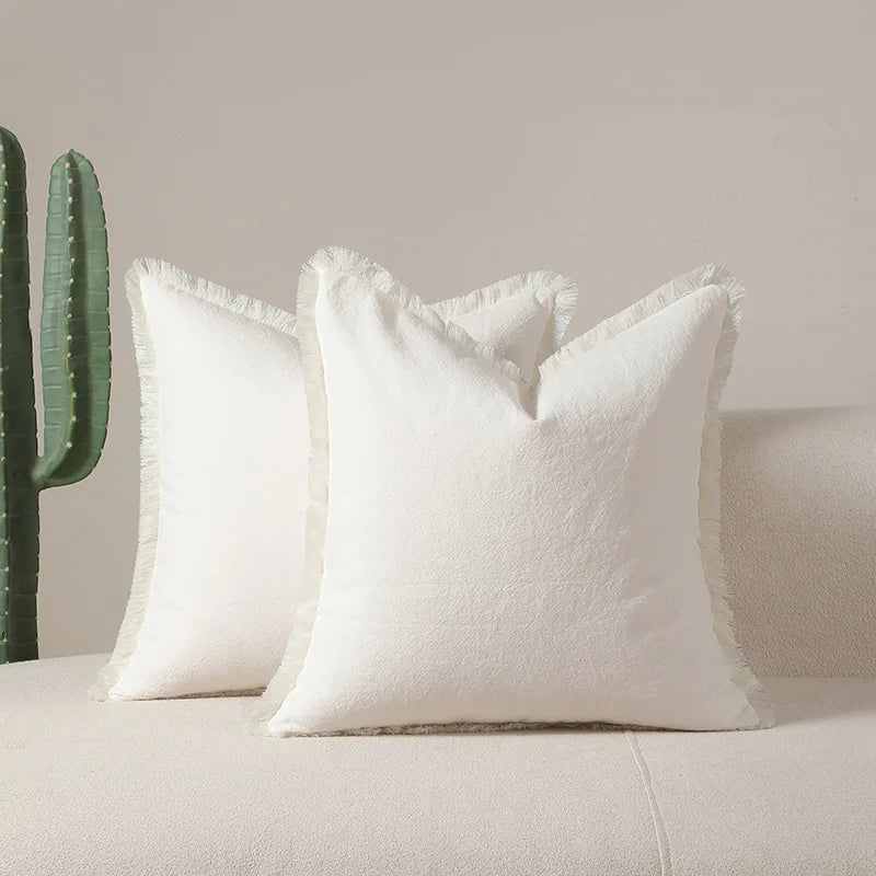 Deux oreillers blancs moelleux avec des franges se dressent devant un fond beige et deux cactus verts.