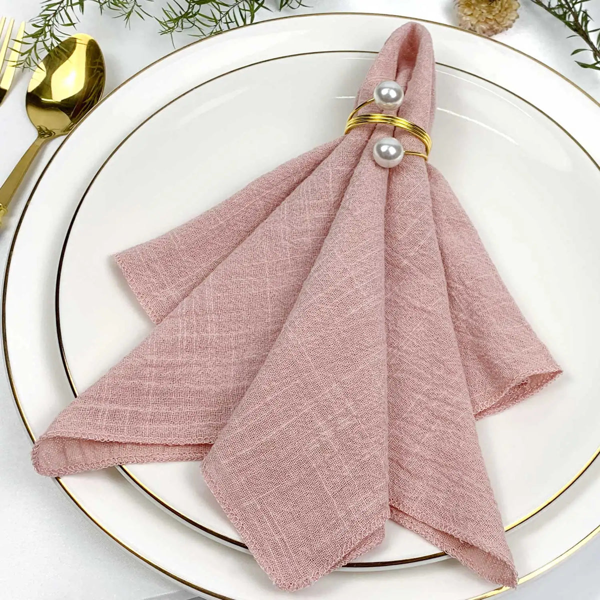 Composition élégante avec une serviette rose pâle pliée de manière décorative sur une assiette blanche avec des accessoires dorés.
