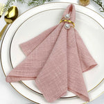 Composition élégante avec une serviette rose pâle pliée de manière décorative sur une assiette blanche avec des accessoires dorés.