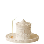 Figurine blanche ornementée d'un carrousel miniature, représentant un château ou une forme architecturale élégante, posée sur un plateau rond.
