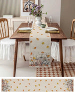 Une table à manger élégante avec une nappe aux motifs de feuilles d'automne, des assiettes blanches et des couverts dorés, le tout sur un fond de meuble en bois et de carrelage à motifs géométriques.