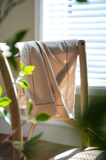 Une couverture en laine beige posée sur une chaise en bois, avec des feuilles vertes apportant une touche naturelle.