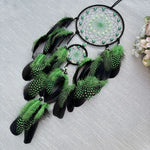 Un joli capteur de rêves aux tons verts et noirs, orné de plumes et de perles, créant une atmosphère mystique et apaisante.