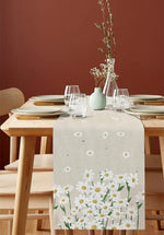 Une table élégante avec une nappe blanche à motifs de marguerites, entourée de vaisselle et de verres sur un fond brun, créant une ambiance chaleureuse.