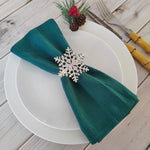 Sur une assiette blanche posée sur un fond en bois clair, une serviette de table verte est pliée et ornée d'un élégant cristal en forme de flocon de neige, avec des cônes de pin et des baies rouges ajoutés comme touche festive.