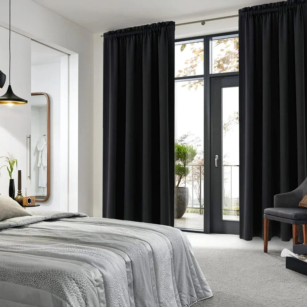 Rideaux noirs occultants posés dans une chambre moderne avec lit gris, baie vitrée et accès à un balcon verdoyant.