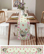 Cette image montre une table de salle à manger joliment décorée avec des fleurs, des assiettes et des verres. Un chemin de table en tissu imprimé de motifs floraux et de lapins de Pâques complète l'ensemble, créant une atmosphère printanière et chaleureuse dans cette pièce.
