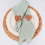 Assiette en céramique blanche avec serviettes de table vertes et accessoires décoratifs en forme de cœur orange sur un napperon tressé de couleur naturelle.
