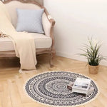 Tapis rond ornemental de style bohème avec motifs ethniques noirs et blancs, placé sur le sol à côté d'un fauteuil vintage et d'un pot de plante verte.