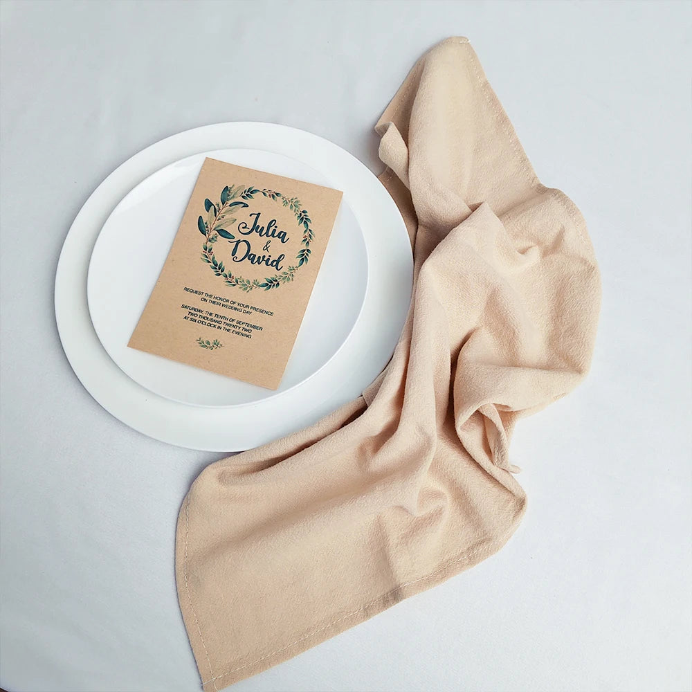Un carton d'invitation avec un motif végétal sur fond blanc, enveloppé dans un tissu beige, sur un plateau blanc.