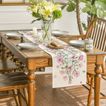 Une table de cuisine en bois décorée d'un bouquet de fleurs jaunes et d'un chemin de table à motifs floraux, avec de la vaisselle et des accessoires pour une atmosphère chaleureuse et accueillante.