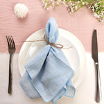 Assiette blanche avec une serviette de table en lin bleu clair, attachée avec une ficelle, sur un fond rose pâle avec des fleurs blanches.