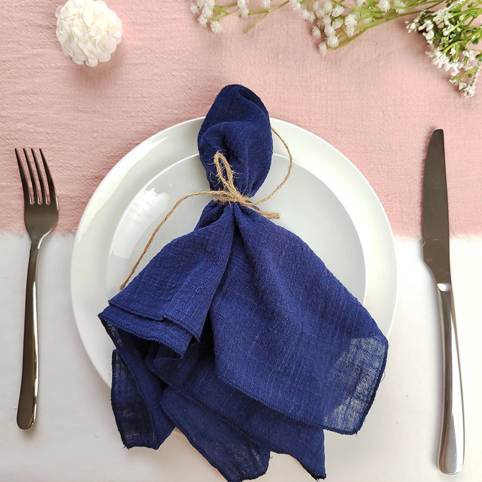Une magnifique serviette en lin bleu marine enroulée avec un brin de ficelle est posée sur une assiette blanche, complétée par des fleurs blanches fraîches sur une surface rose clair.