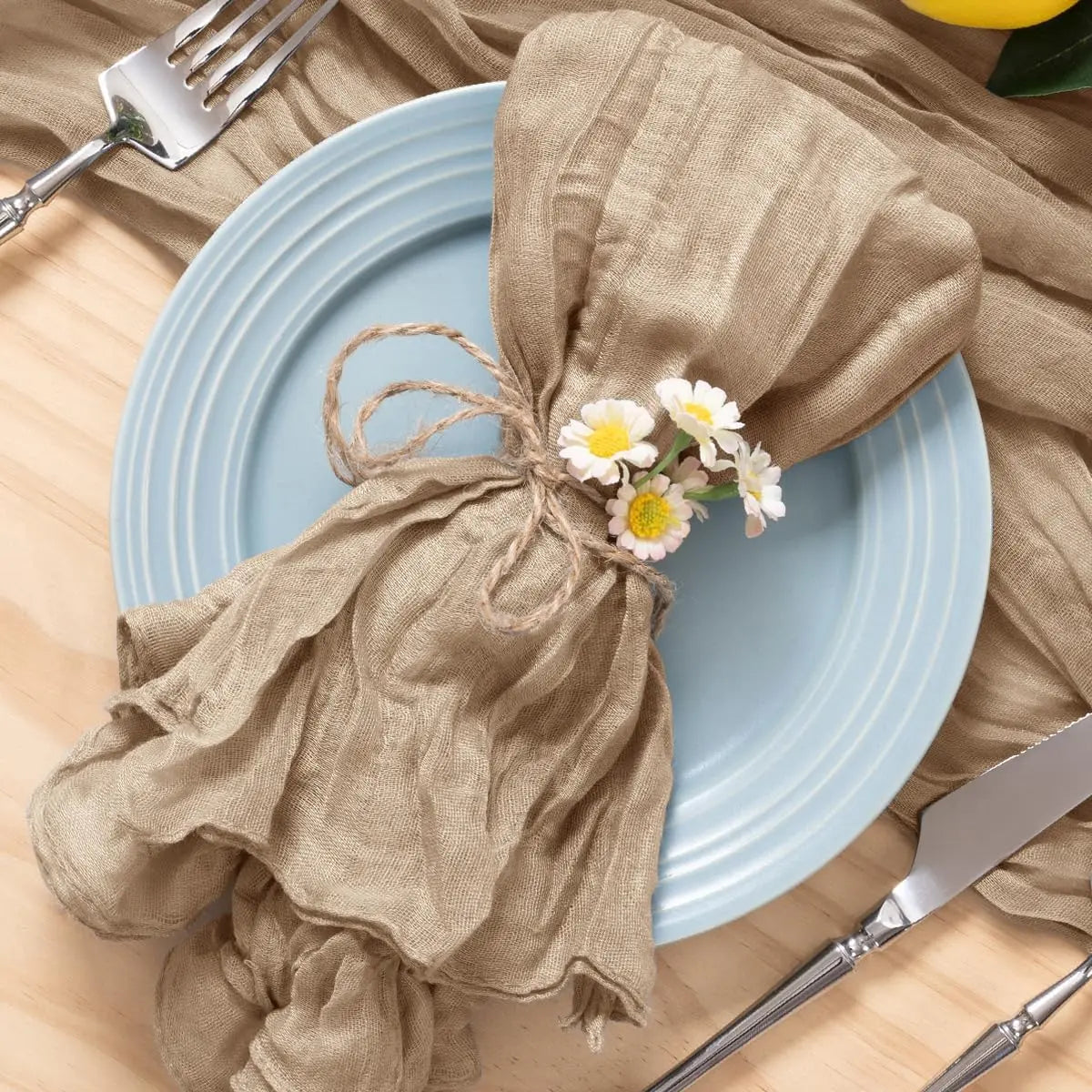 Une assiette bleue sur une table en bois, avec des couverts et un linge de table en lin brun orné de fleurs de marguerite, créant une ambiance rustique et naturelle.