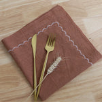 Une serviette en tissu marron, une fourchette et un couteau en métal doré, accompagnés d'épis de blé, créent une composition élégante et naturelle sur un fond en bois clair.