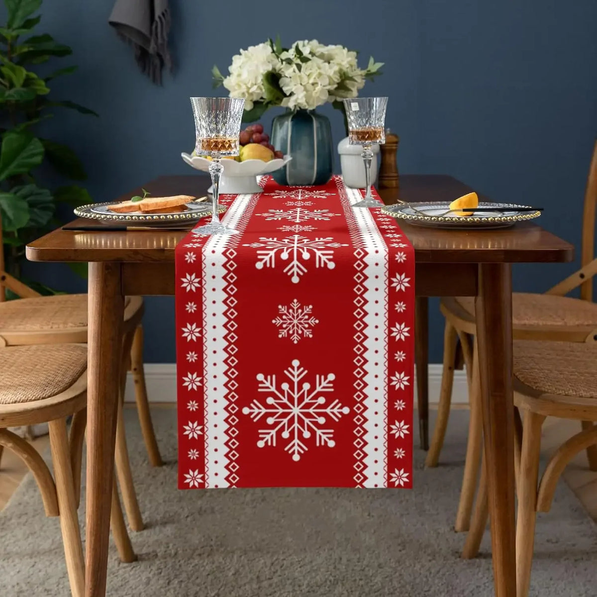 Table décorée dans un style hivernal avec nappe rouge ornée de motifs de flocons de neige blancs, vases de fleurs blanches, verres de cristal et assiettes sur une table en bois.