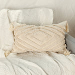 Cette image montre un coussin décoratif moelleux et texturé avec des motifs géométriques et des franges sur un tissu blanc crème, créant une ambiance douce et relaxante dans un intérieur.