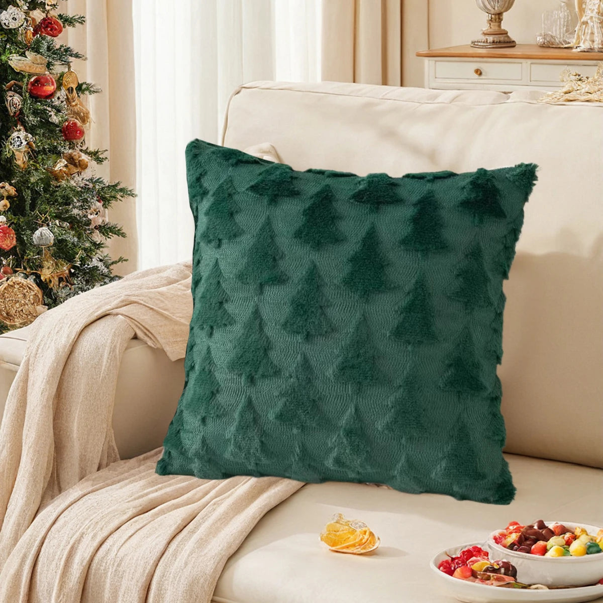 Un coussin décoratif vert foncé orné d'un motif de sapin, placé sur un canapé beige dans un salon décoré pour Noël avec un arbre de Noël et des plats de fruits sur une table.