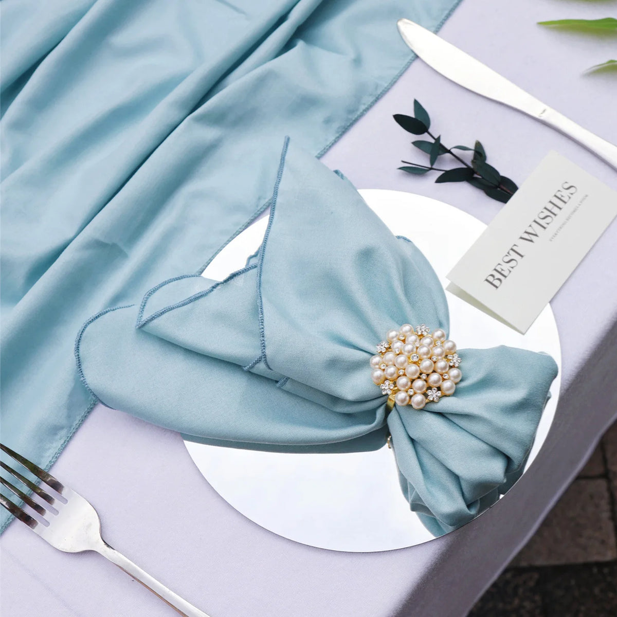 Une serviette de table bleu clair avec une broche ornée de perles, entourée d'une carte avec le logo d'une marque de décoration.