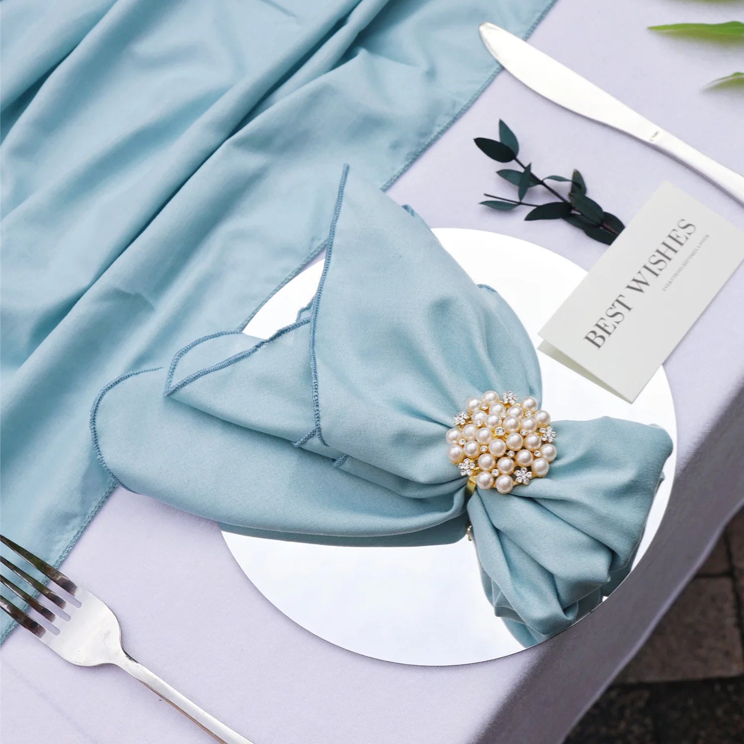 Une serviette de table bleu clair avec une broche ornée de perles, entourée d'une carte avec le logo d'une marque de décoration.