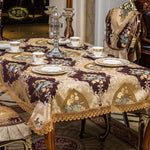 La table est drapée d'une nappe en soie brune et dorée finement ornée de fleurs, entourée de tasses et d'assiettes en porcelaine, ainsi que d'un magnifique chandelier en cristal, dans un décor luxueux et raffiné.