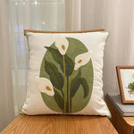Coussin décoratif en tissu beige avec un motif brodé de fleurs de calla blanches sur des feuilles vertes.