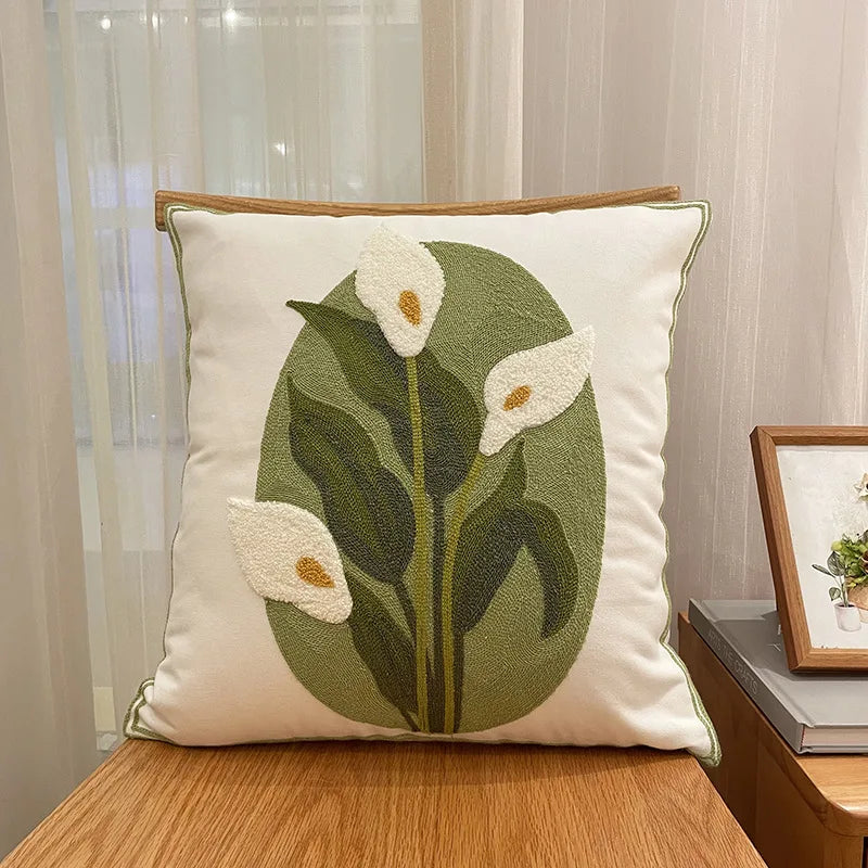 Coussin décoratif en tissu beige avec un motif brodé de fleurs de calla blanches sur des feuilles vertes.