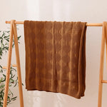 Une couverture tricotée en motif géométrique de couleur marron posée sur un support en bois, avec des éléments verts en arrière-plan.