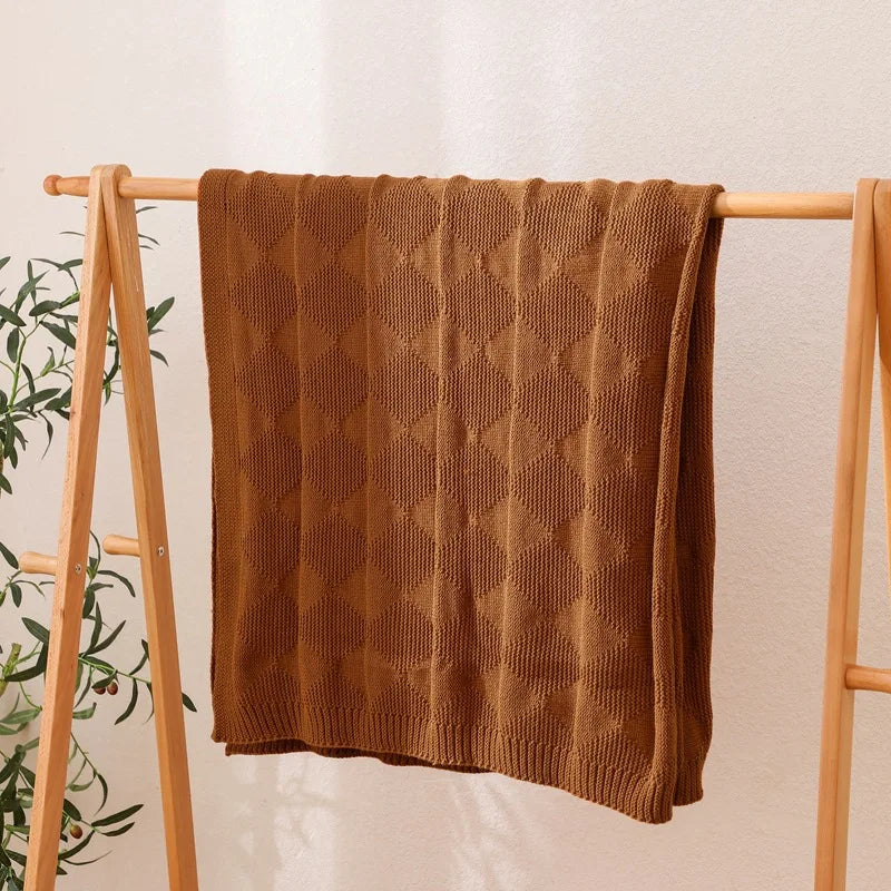 Une couverture tricotée en motif géométrique de couleur marron posée sur un support en bois, avec des éléments verts en arrière-plan.
