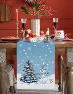 Une table de Noël joliment décorée avec un centre de table festif, des verres à vin et une bougie allumée, rehaussée par un chemin de table décoré d'un motif de flocons de neige et d'un bonhomme de neige.