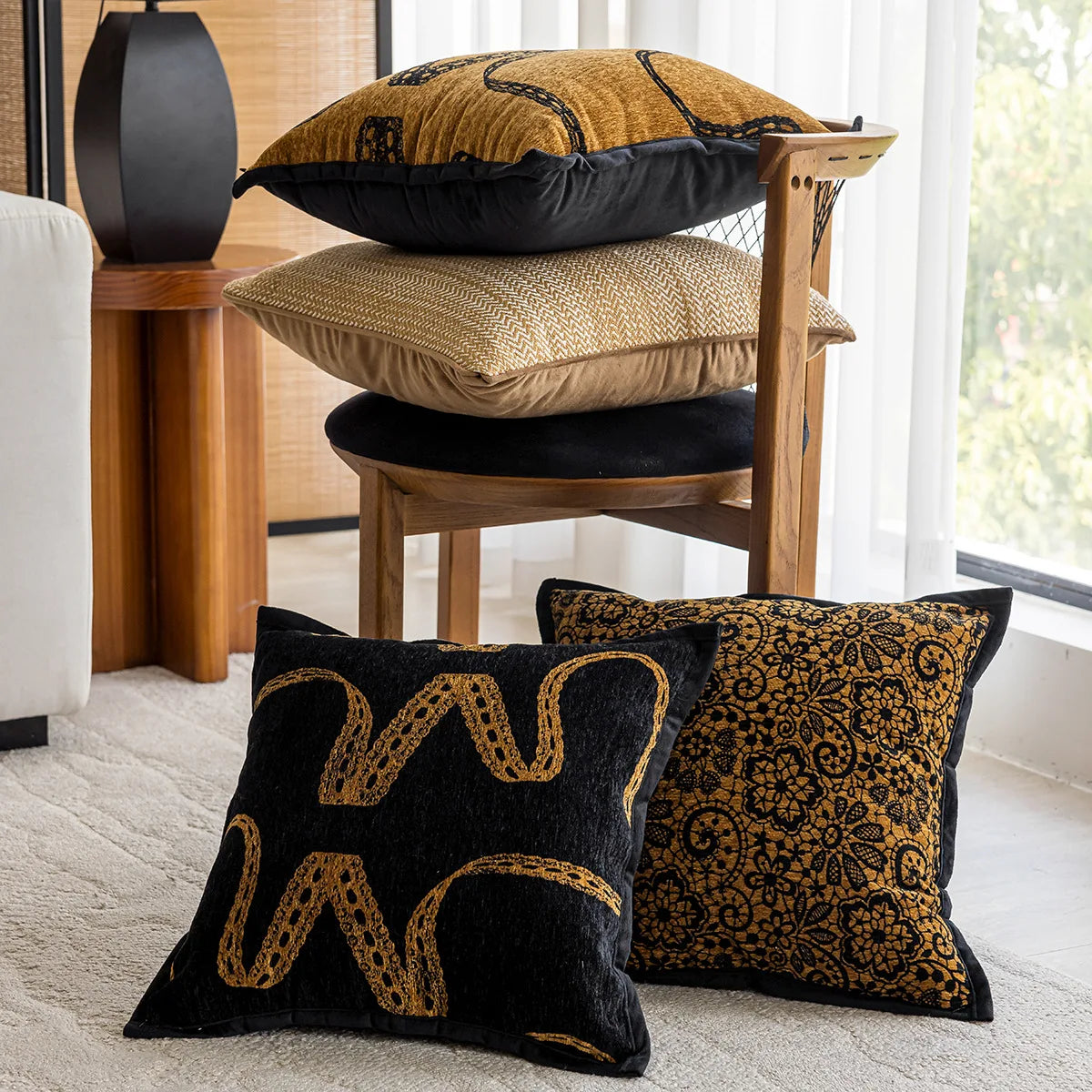 Coussins décoratifs en velours noir et doré avec motifs floraux et abstraits, disposés sur un meuble en bois avec d'autres coussins et éléments de décoration dans un intérieur moderne et lumineux.