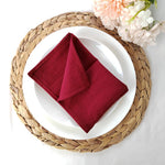 Une jolie nappe de table tressée sert de support à une assiette blanche avec une serviette en tissu rouge bordeau pliée au centre, accompagnée de fleurs colorées.