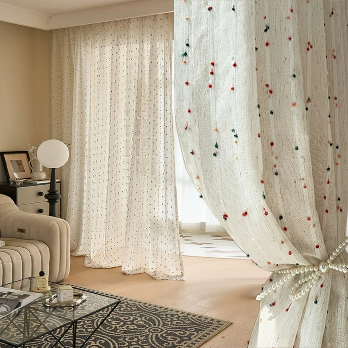 Voilage crème à pois multicolores tiré sur le côté avec embrasse perle, dans un salon cosy aux tons beige et bois.