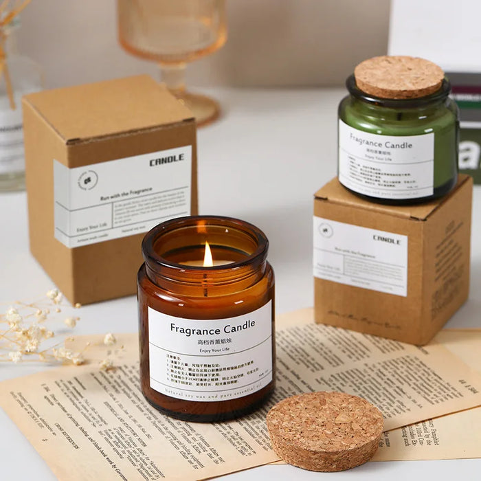Bougie parfumée dans un bocal en verre ambré, accompagnée de boîtes en carton portant l'étiquette "Fragrance Candle", sur un journal ouvert.