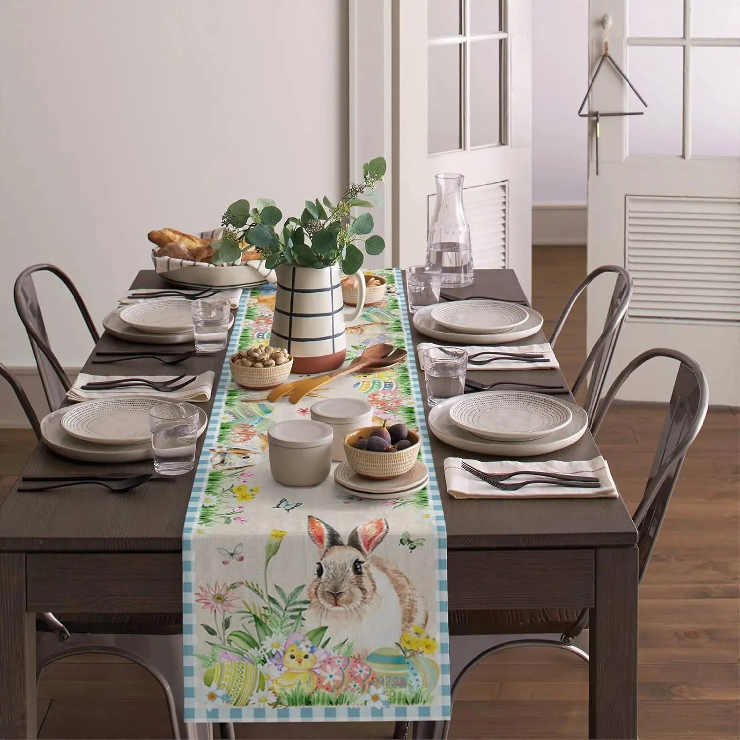 Une table de salle à manger élégante avec un chemin de table décoré de motifs printaniers et d'un lapin mignon, accompagné de vaisselle blanche et de plantes vertes, créant une ambiance chaleureuse et naturelle.