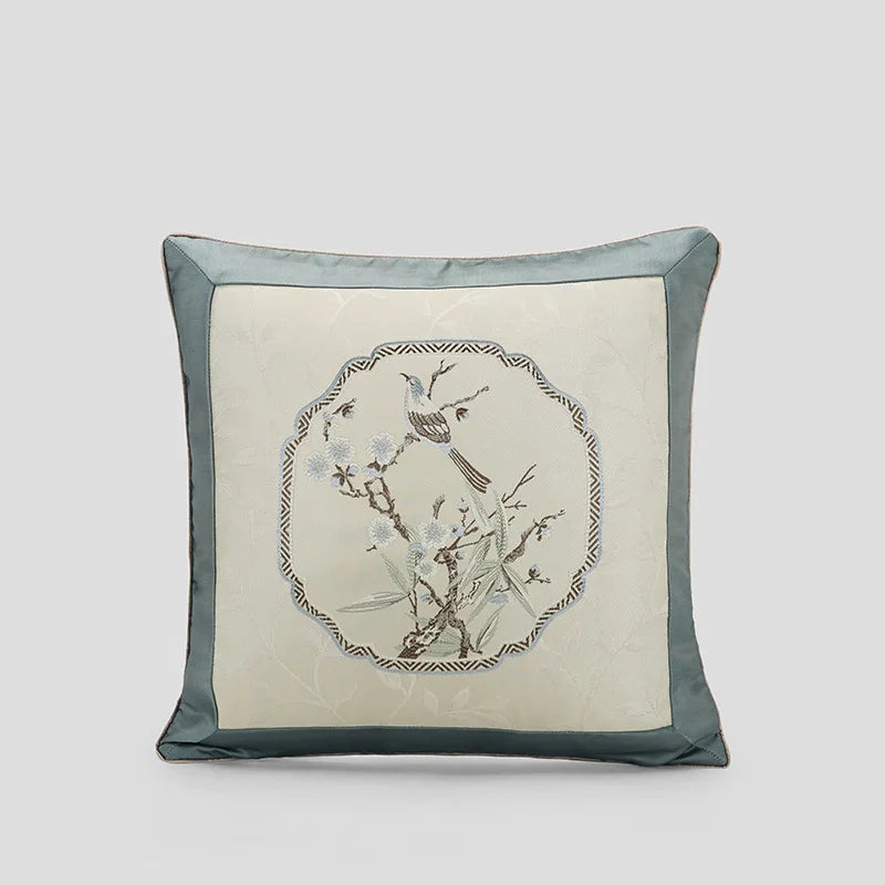 Coussin décoratif avec motif de branche fleurie et oiseau brodé dans un cadre géométrique sur fond ivoire et gris.