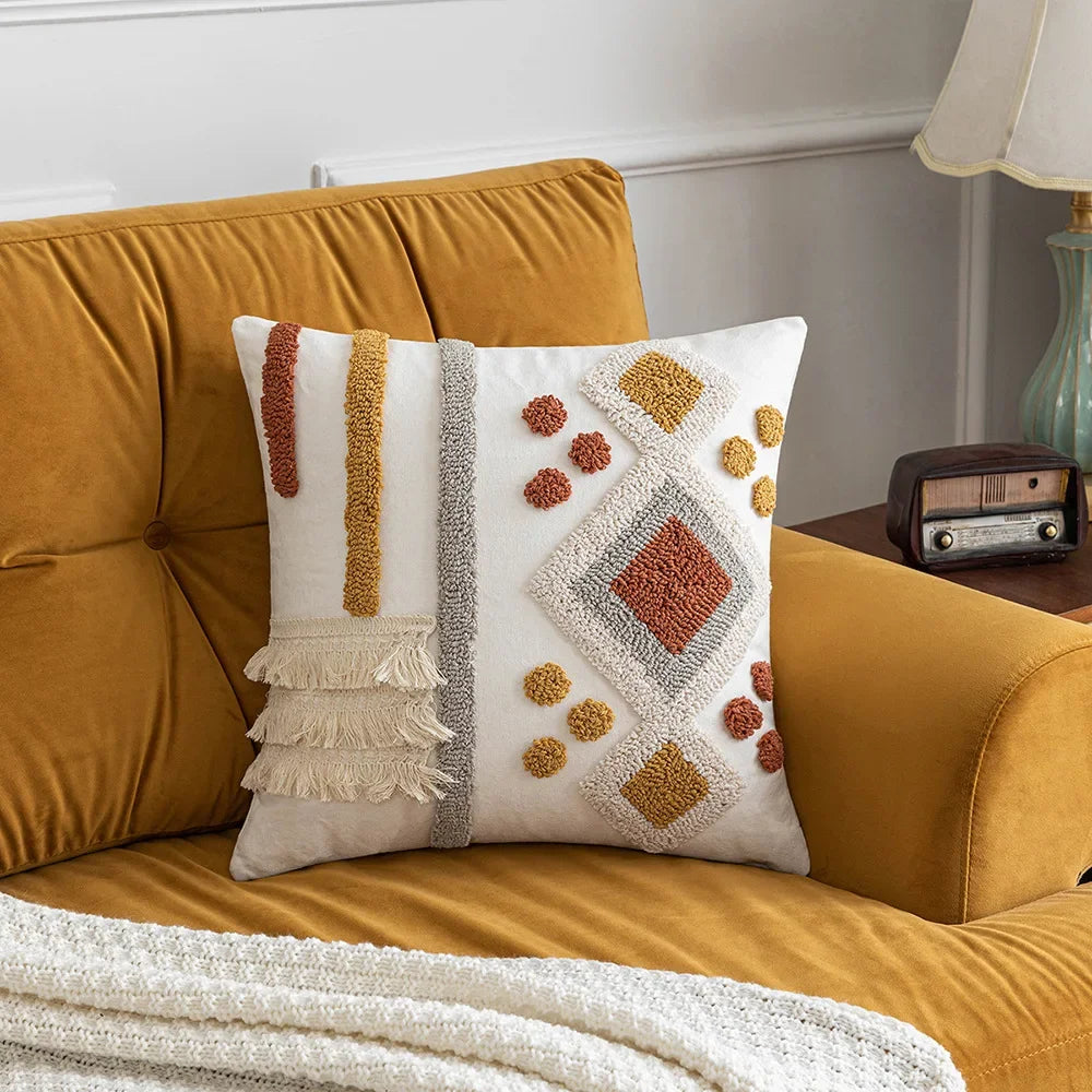 Un oreiller décoratif avec des motifs géométriques et des pompons dans des tons de beige, jaune et marron posé sur un canapé jaune dans un intérieur vintage.