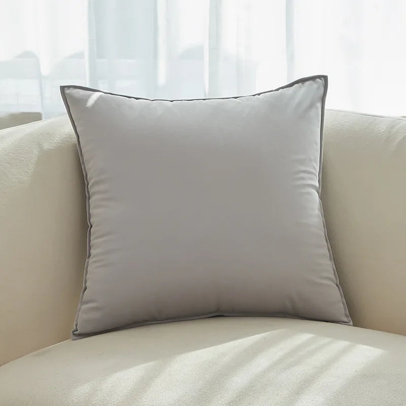 Coussin décoratif gris clair sur un canapé confortable au premier plan, avec un intérieur lumineux et des rideaux blancs en arrière-plan.