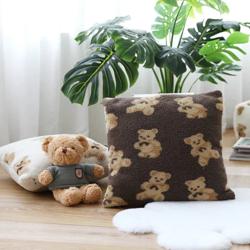 Un coussin moelleux à motifs d'ours en peluche et une figurine de peluche posés sur une table en bois, avec des plantes vertes en arrière-plan.