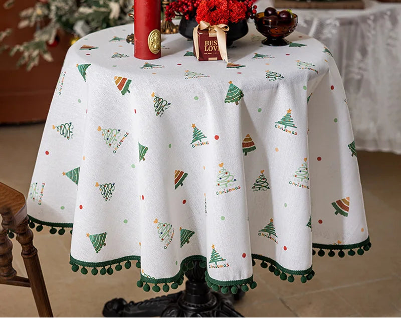 Une nappe de table ronde blanche ornée de motifs d'arbres de Noël et de pompons vert émeraude, avec au centre un bouquet de fleurs rouges, une bougie rouge et une décoration cadeau, créant une ambiance festive de Noël.