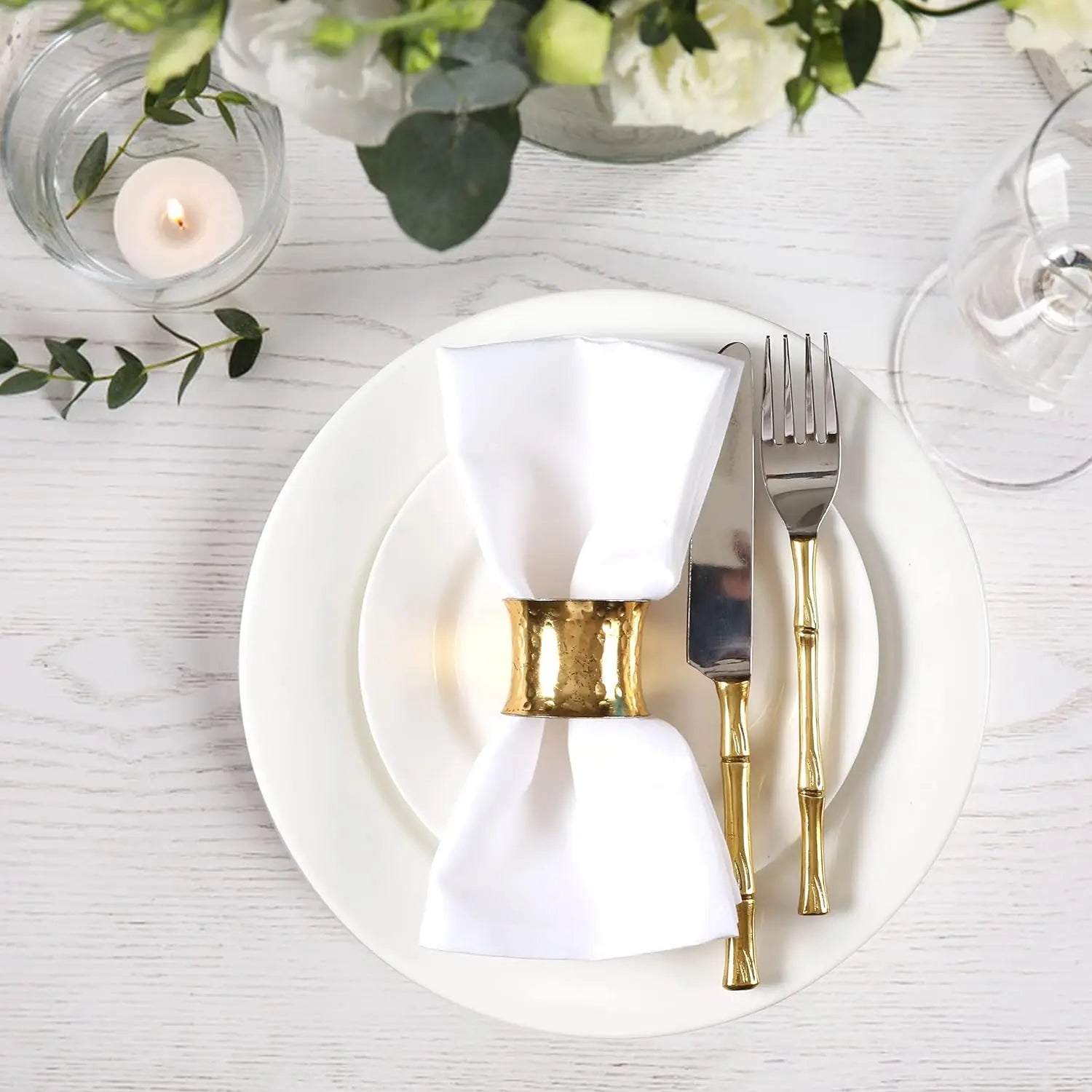 Un beau cadre élégant avec une assiette blanche, des couverts dorés, une serviette blanche enroulée dans un anneau doré, et une décoration florale sur une table en bois blanc.