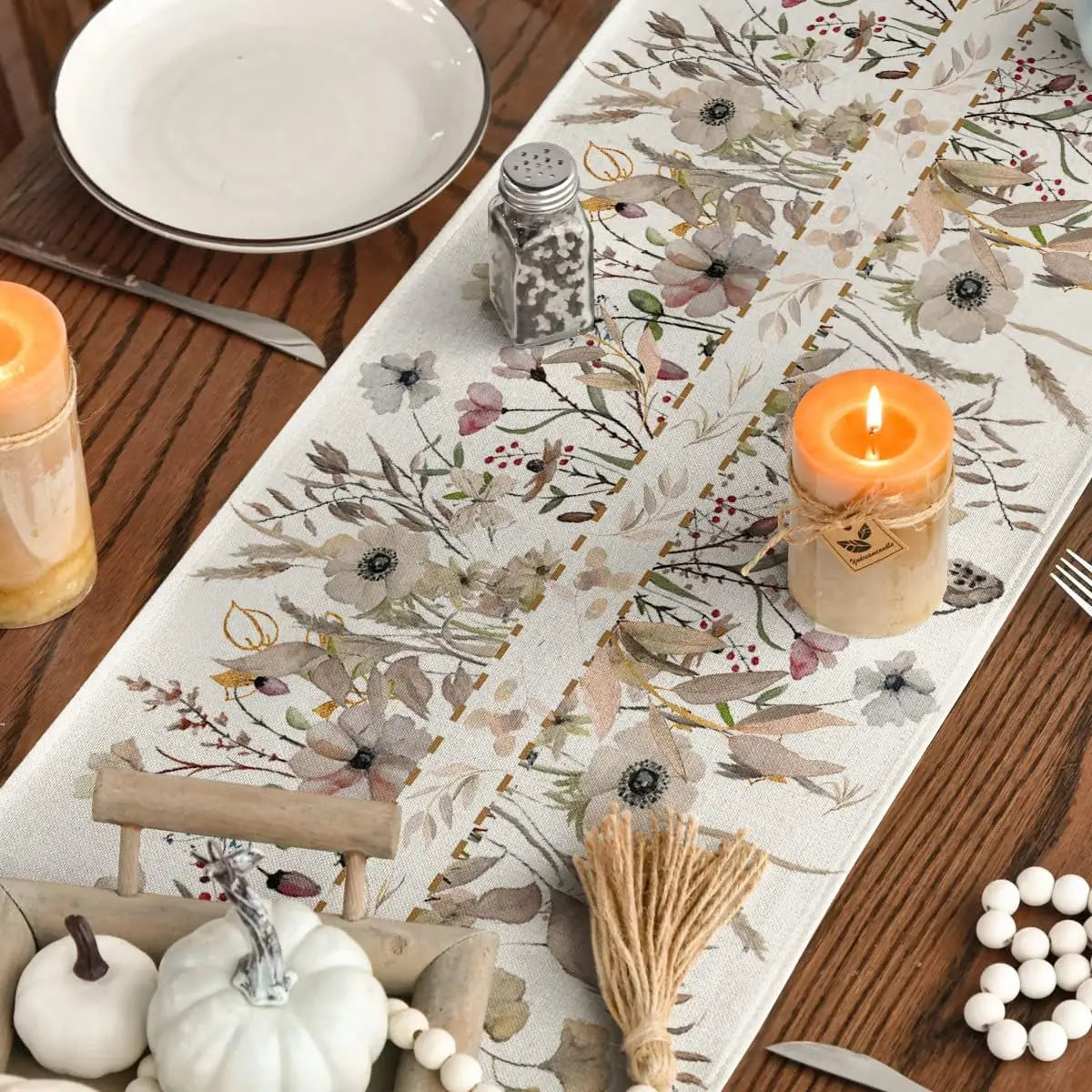 Décor de table automnal avec nappe florale, bougie allumée et divers accessoires décoratifs tels que citrouilles et perles.