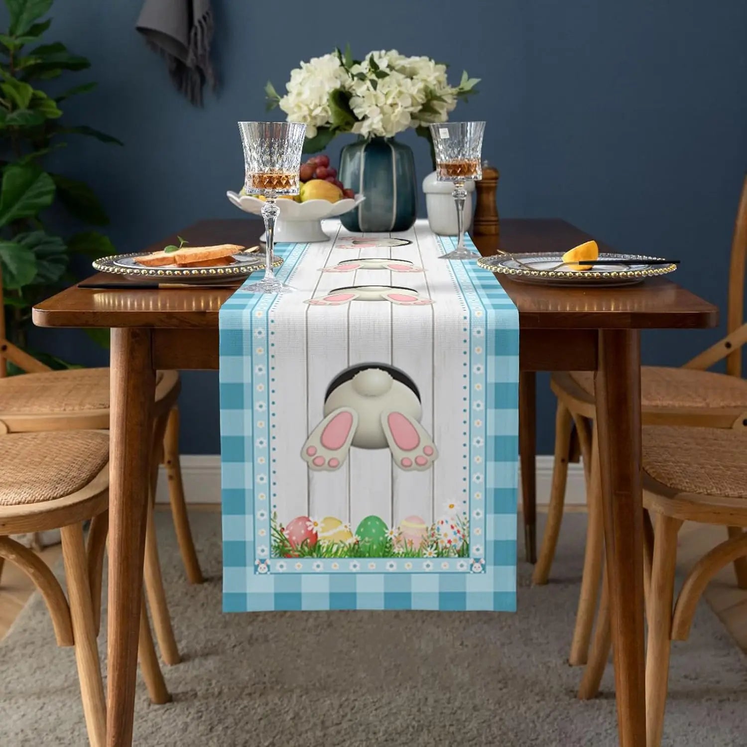 Table de salle à manger avec un chemin de table décoré de motifs de Pâques, incluant des oeufs colorés et un lapin en céramique, accompagné de fleurs et de verres de vin.