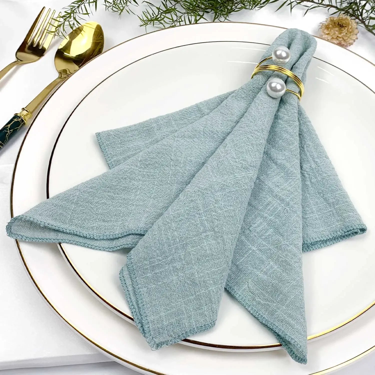 Une serviette en tissu bleu clair est élégamment posée sur une assiette blanche avec une fourchette et un couteau dorés.