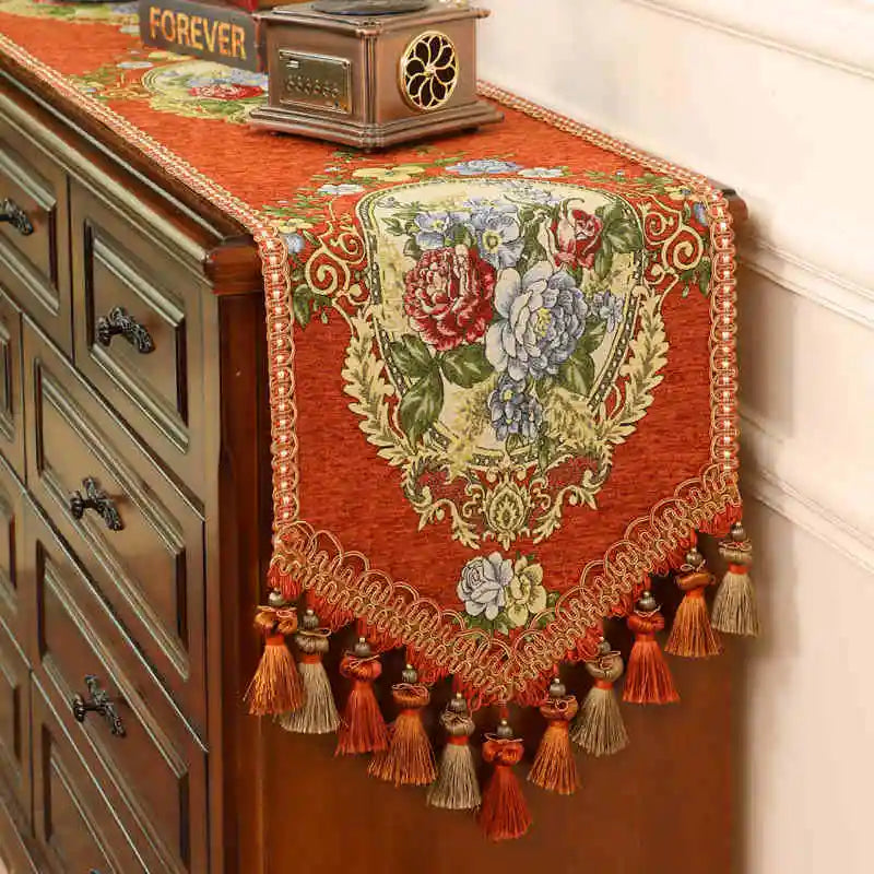 Un superbe chemin de table en tissu rouge orné de fleurs aux couleurs vives, avec des pompons et des glands décoratifs qui lui donnent un charme vintage.