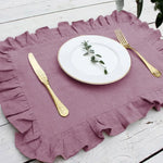 Une serviette en lin violet ornée de volants, accompagnée d'une assiette blanche et de couverts dorés, évoquant une ambiance élégante et raffinée.